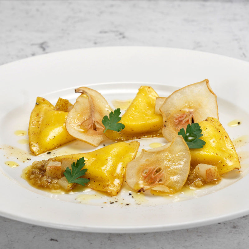 Four pieces of Divine Creazioni Triangoletti with Parmigiano Reggiano DOP are presented on a white plate with pear slices, small cubes, parsley leaves, light sauce, and a garnish of cracked black pepper.