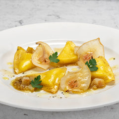 Four pieces of Divine Creazioni Triangoletti with Parmigiano Reggiano DOP are presented on a white plate with pear slices, small cubes, parsley leaves, light sauce, and a garnish of cracked black pepper.