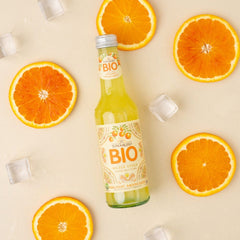 A bottle of Tomarchio Organic Aranciata Orange Soda DOP (24-Pack, 275ml) sits among fresh orange slices and ice cubes on a light beige backdrop, highlighting the refreshing taste of authentic Sicilian orange soda.