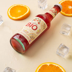 A bottle of Tomarchio Organic Blood Orange Soda IGP (275ml) from a 24-pack rests on a light surface with orange slices and ice cubes, condensation beading on the glass. Made with Arancia Rossa di Sicilia IGP blood oranges.